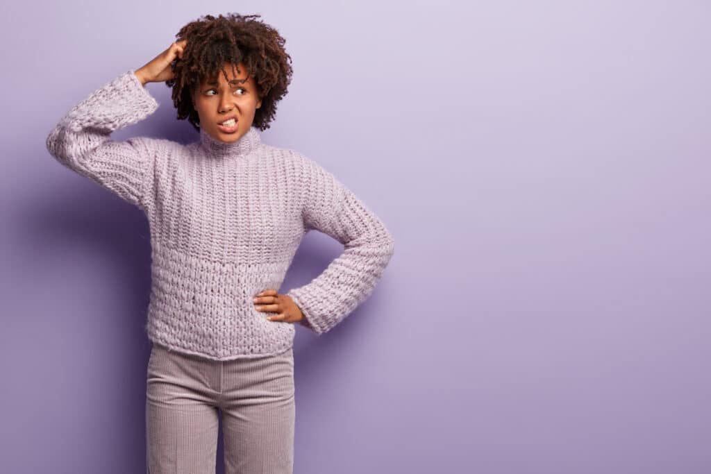 Unsure black young woman scatches back of head, looks aside with confused unaware expression, clenches teeth, wears jumper and trousers, keeps hand on waist, thinks doubtfully, makes decision