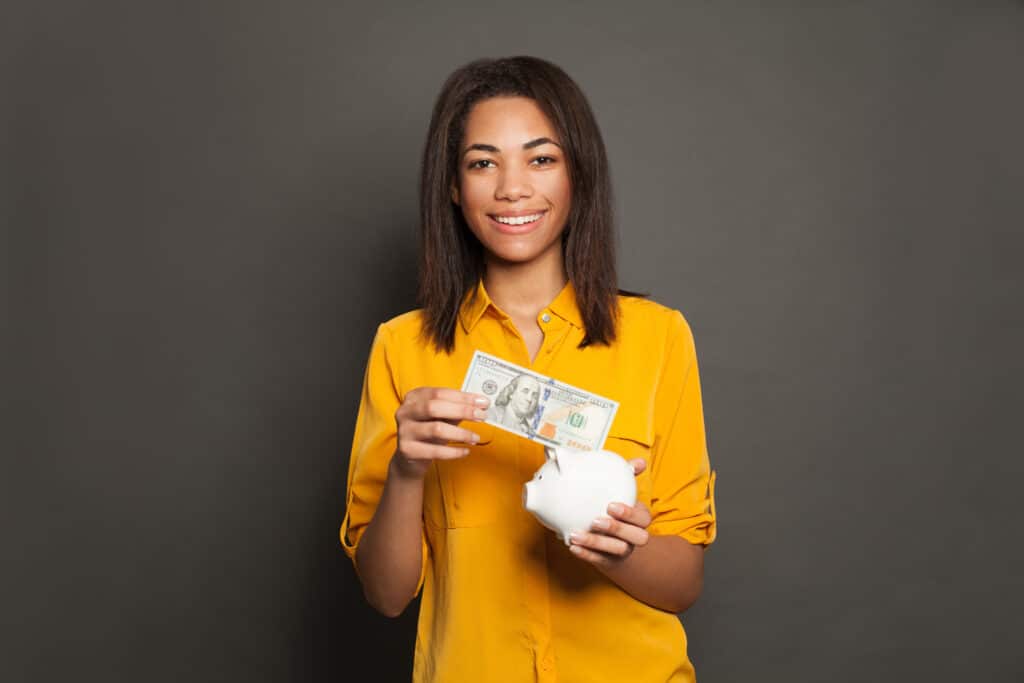 Smart happy successful black woman putting in piggy bank one hundred dollars banknote