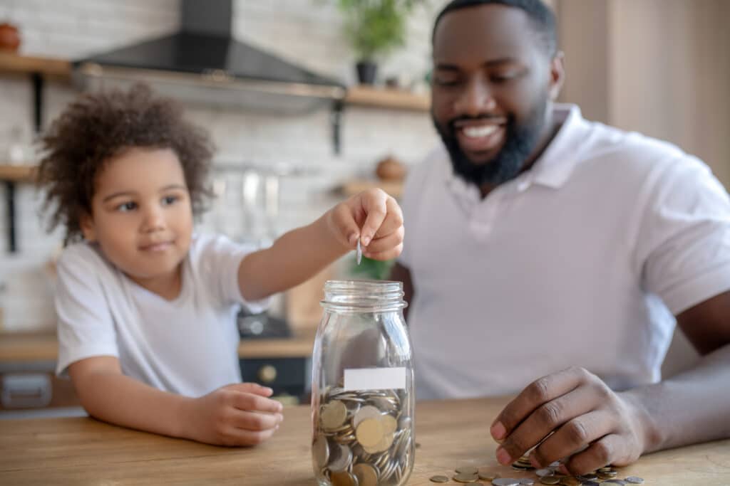Putting coins inside. Cute curly-haired kid putting coins into the moneybank representing a custodial roth ira