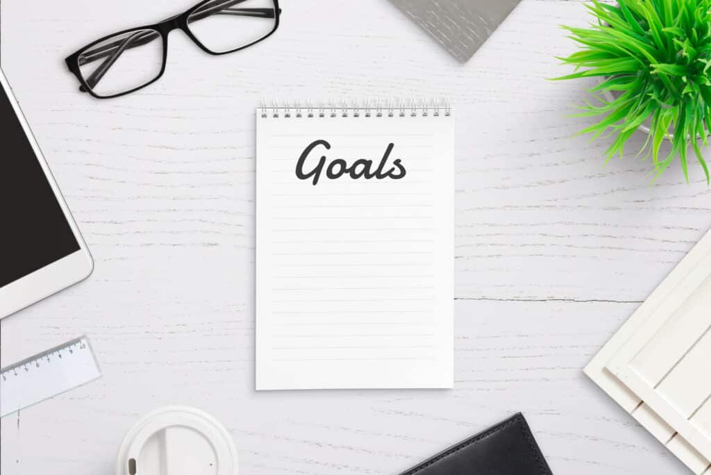 Writing goals on empty notepad page with goals title in header. Top view office desk with smart phone, cup of coffee, glasses, plant representing smart goals examples for work