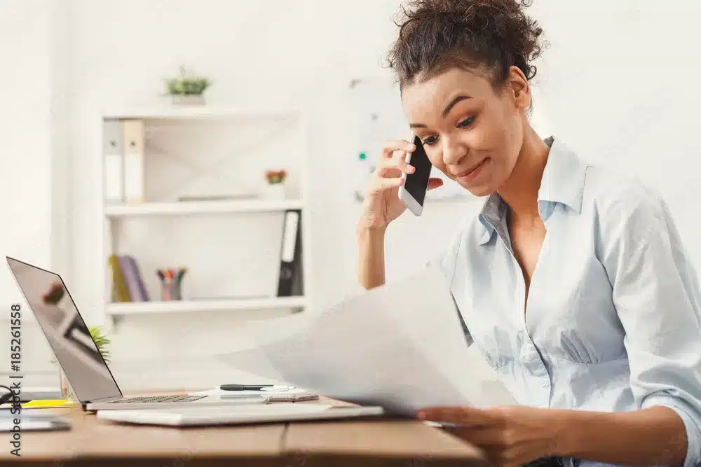 A woman seated at her desk with a laptop, talking on the phone, and holding a paper about 'How to Buy IPOs'.