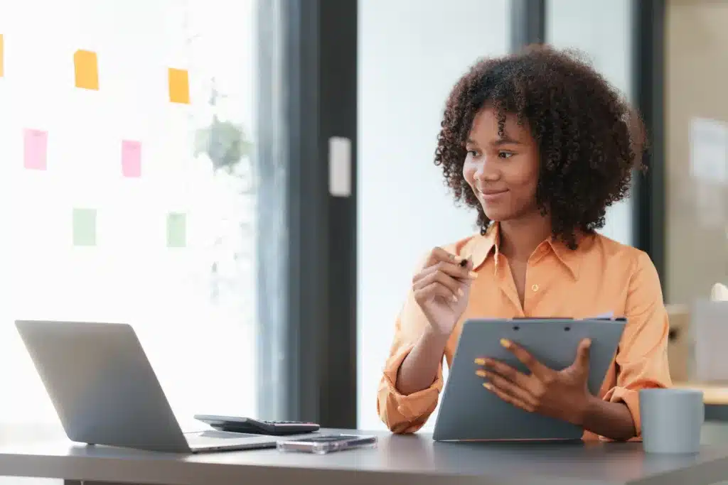 A woman sits at her office desk, attentively reviewing notes spread out in front of her laptop while focusing on the topic of Regular Financial Review and Analysis.