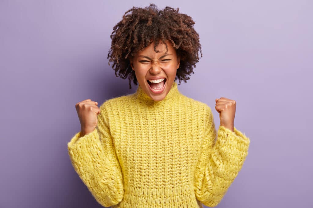 Horizontal shot of overjoyed glad woman with curly hair, clenches fists like winner, exclaims positively, sure in success, celebrates achieved success, wears yellow winter jumper, feels pleasure